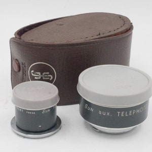 May include: Two vintage camera lens attachments in a brown leather case. The lenses are labeled "Sun aux. TELEPHOTO" and "Sun 1:2.8 50mm 1/487 Japan".