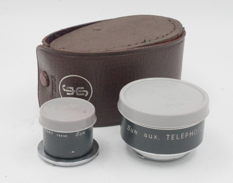 May include: Two vintage camera lens attachments in a brown leather case. The lenses are labeled "Sun aux. TELEPHOTO" and "Sun 1:2.8 50mm 1/487 Japan".