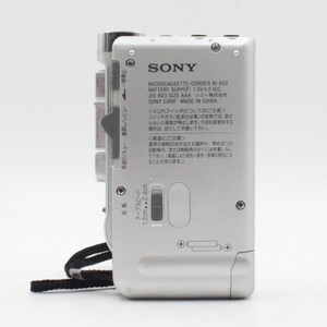 May include: A silver Sony Microcassette-Corder M-850 with a black strap. The device has a tape speed switch, a volume knob, and a battery compartment. The text on the back reads "SONY Microcassette-Corder M-850 Battery Supply: 1.5V x 2 IEC, JIS R03 SIZE AAA ソニー株式会社 SONY CORP. MADE IN CHINA".