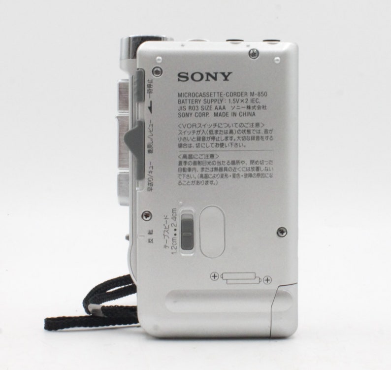 May include: A silver Sony Microcassette-Corder M-850 with a black strap. The device has a tape speed switch, a volume knob, and a battery compartment. The text on the back reads "SONY Microcassette-Corder M-850 Battery Supply: 1.5V x 2 IEC, JIS R03 SIZE AAA ソニー株式会社 SONY CORP. MADE IN CHINA".