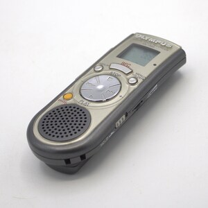 May include: A silver Olympus digital voice recorder with a black speaker and a circular button with the words "PLAY" and "STOP" printed on it. The recorder has a small LCD screen with the word "REC" displayed on it.