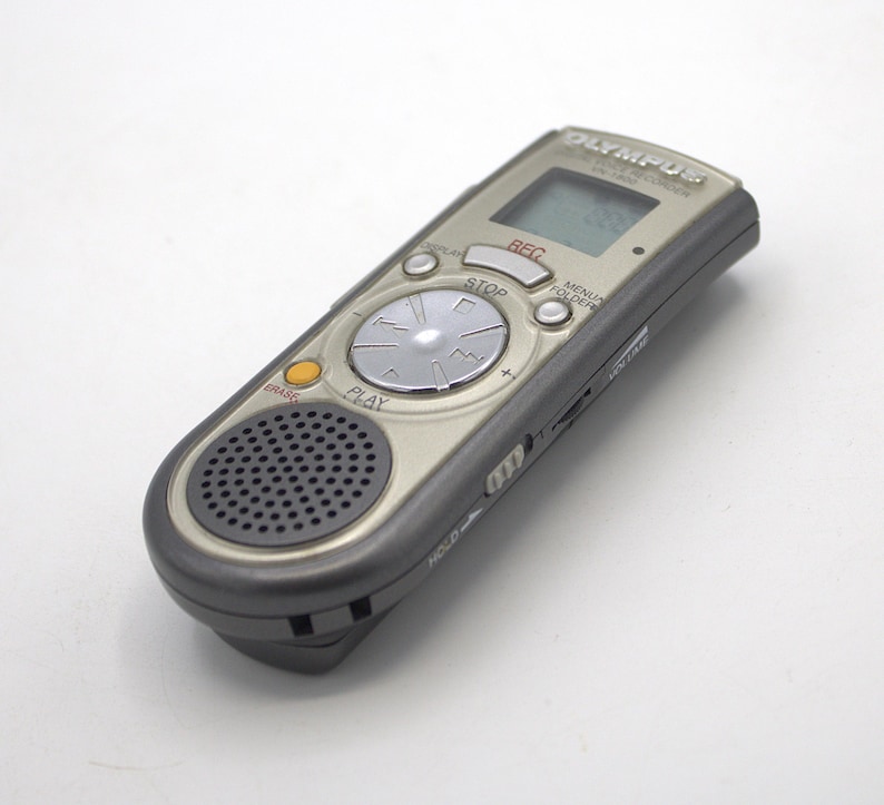 May include: A silver Olympus digital voice recorder with a black speaker and a circular button with the words "PLAY" and "STOP" printed on it. The recorder has a small LCD screen with the word "REC" displayed on it.