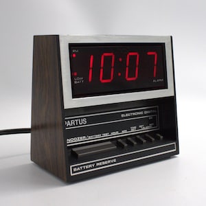 May include: A vintage Partus digital alarm clock with a wood-grain finish. The red LED display shows 10:07 PM. Features include a snooze button and battery reserve. The clock is black and silver.