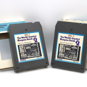 May include: Two vintage 8-track tapes, "The World's Greatest Bluegrass Bands," Volumes 1 and 2, with their original boxes. The tapes are dark gray with colorful labels. The boxes are white and blue, with the Columbia House logo.