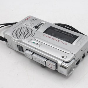 May include: A silver Sony voice recorder with a black strap. The recorder has a built-in microphone and a clear voice plus feature. The text "AUTO REVERSE" is visible on the recorder.