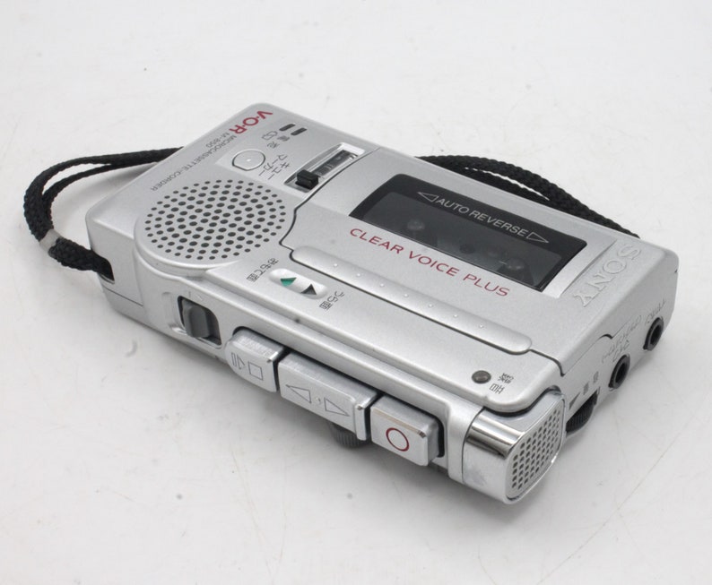 May include: A silver Sony voice recorder with a black strap. The recorder has a built-in microphone and a clear voice plus feature. The text "AUTO REVERSE" is visible on the recorder.