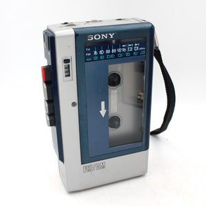 May include: A vintage Sony FM/AM cassette player in a silver and blue casing. The player has a clear window to view the cassette tape and a black carrying strap. The radio dial is visible, and the buttons are red and black.