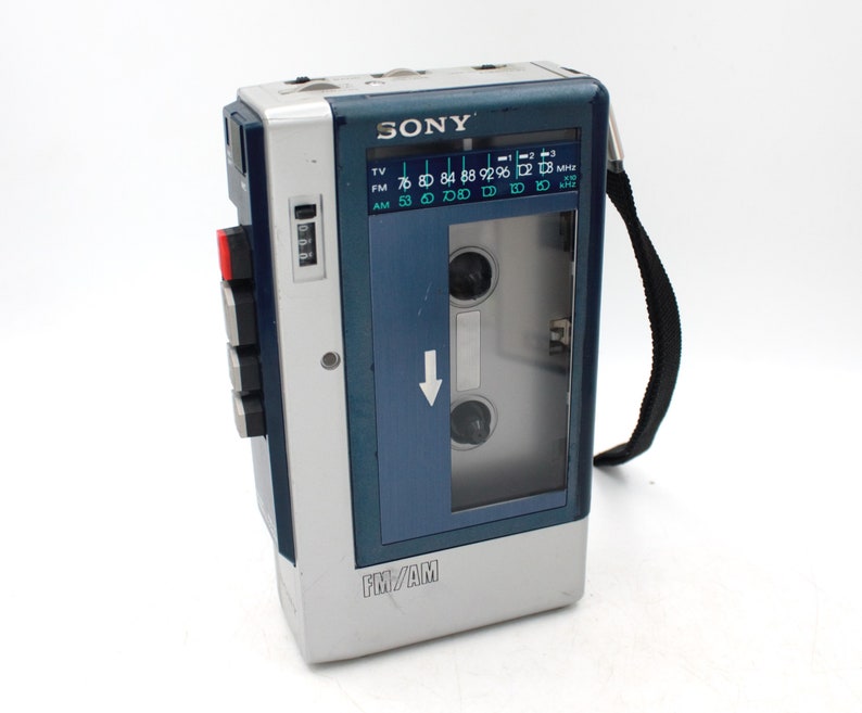 May include: A vintage Sony FM/AM cassette player in a silver and blue casing. The player has a clear window to view the cassette tape and a black carrying strap. The radio dial is visible, and the buttons are red and black.