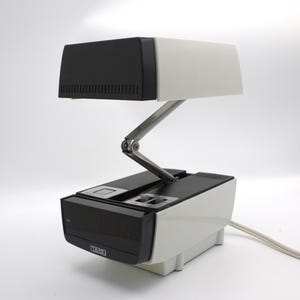 May include: A vintage Cosmo desk lamp with a black and white design. The lamp features an adjustable arm and a rectangular shade. The base has a digital clock display and the word "COSMO" is printed on the front.