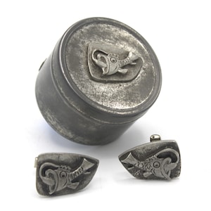 Puede incluir: Un juego de gemelos de plata y una pequeña caja redonda con un diseño de pez. Los gemelos y la caja tienen un aspecto desgastado y envejecido.