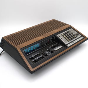 May include: A General Electric programmable clock radio with a wood grain finish and black plastic casing. The device features a digital display showing 12:02, a numeric keypad, sliding volume and tone controls, and various function switches.