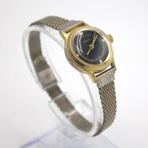 May include: A vintage Geneva wristwatch with a gold-tone case and a black dial. The watch features a mesh metal band and a small, round face with gold hands and hour markers. The watch is displayed on a clear stand.