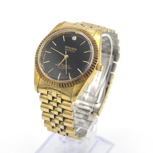 May include: A gold-tone Gruen Precision watch with a black face and a diamond at the 12 o'clock position. The watch has a gold-tone metal band and says "3 ATM-K03T WATER RESISTANT QUARTZ" on the face.