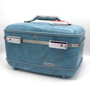 May include: A vintage blue Tourister suitcase with a silver handle and trim. The suitcase has a textured surface, metal latches, and a small tag that reads "American Tourister". The Tourister logo is visible on the front.