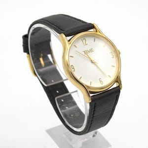 May include: A gold-toned wristwatch with a white face and black leather band. The watch face displays the word "TIME" and the numbers 3, 6, 9, and 12. The watch is displayed on a clear stand.