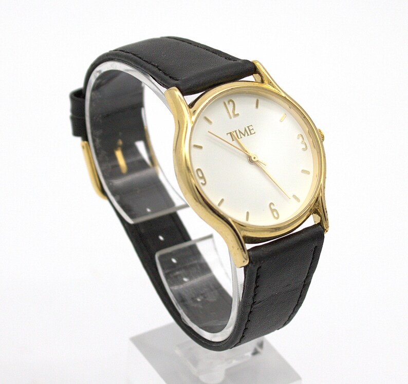 May include: A gold-toned wristwatch with a white face and black leather band. The watch face displays the word "TIME" and the numbers 3, 6, 9, and 12. The watch is displayed on a clear stand.