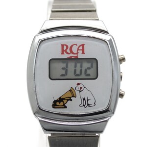 May include: A silver-tone digital wristwatch with a white face featuring the red RCA logo, a digital time display showing 3:02, and a graphic of a dog sitting next to a gramophone. The watch has a metallic link band and three side buttons.