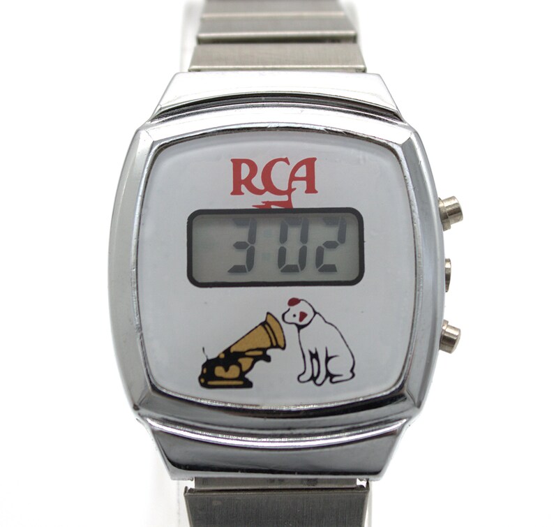 May include: A silver-tone digital wristwatch with a white face featuring the red RCA logo, a digital time display showing 3:02, and a graphic of a dog sitting next to a gramophone. The watch has a metallic link band and three side buttons.
