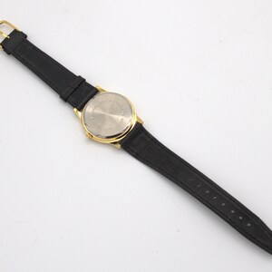 May include: A gold-toned wristwatch with a black leather band. The watch has a round face and a gold buckle. The back of the watch is silver-toned. The watch band is approximately 9 inches long.