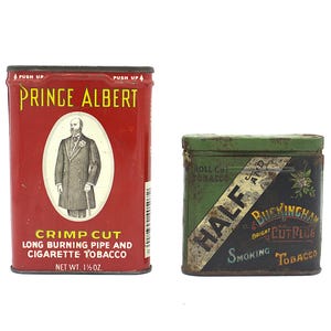 May include: Two vintage tobacco tins. The first tin is red with gold lettering and features a man in a suit and top hat. The text on the tin reads "Prince Albert Crimp Cut Long Burning Pipe and Cigarette Tobacco Net Wt. 1 1/4 oz.". The second tin is green and black with gold lettering and features the text "Roll Cut Tobacco Half and Buckingham Bright Cut Plug Smoking Tobacco".
