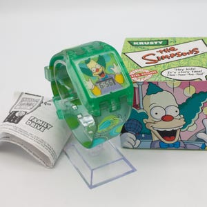 May include: A green Krusty the Clown watch from The Simpsons, with a digital display showing 6:56. The watch is in its original packaging, which features Krusty's image. Includes a Family Drive instruction sheet.