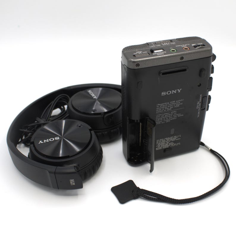 May include: Black Sony headphones and a black Sony digital voice recorder are displayed on a white background. The headphones are folded, and the recorder has a battery compartment open. A black lanyard is attached to the recorder.