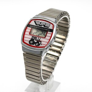Peut inclure: Montre num&eacute;rique vintage avec bracelet extensible en m&eacute;tal argent&eacute;. Le cadran affiche le texte "WHITE FARM EQUIPMENT" et un dessin de tracteur. La montre affiche l'heure. La montre est sur un support transparent.