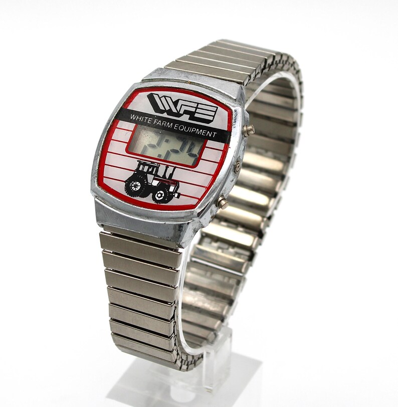 Peut inclure: Montre num&eacute;rique vintage avec bracelet extensible en m&eacute;tal argent&eacute;. Le cadran affiche le texte "WHITE FARM EQUIPMENT" et un dessin de tracteur. La montre affiche l'heure. La montre est sur un support transparent.