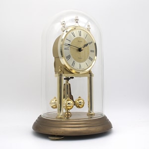 May include: A Kundo quartz anniversary clock with a gold-tone face, black Roman numerals, and black hands. The clock features a rotating pendulum with four gold spheres, enclosed within a clear glass dome on a circular brass-colored base.