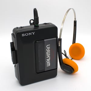 May include: A black Sony Walkman cassette player with a gray center panel and the word Walkman printed in white. A black headphone cord is plugged into the top. Included are black headphones with bright orange foam ear pads and a metal headband.