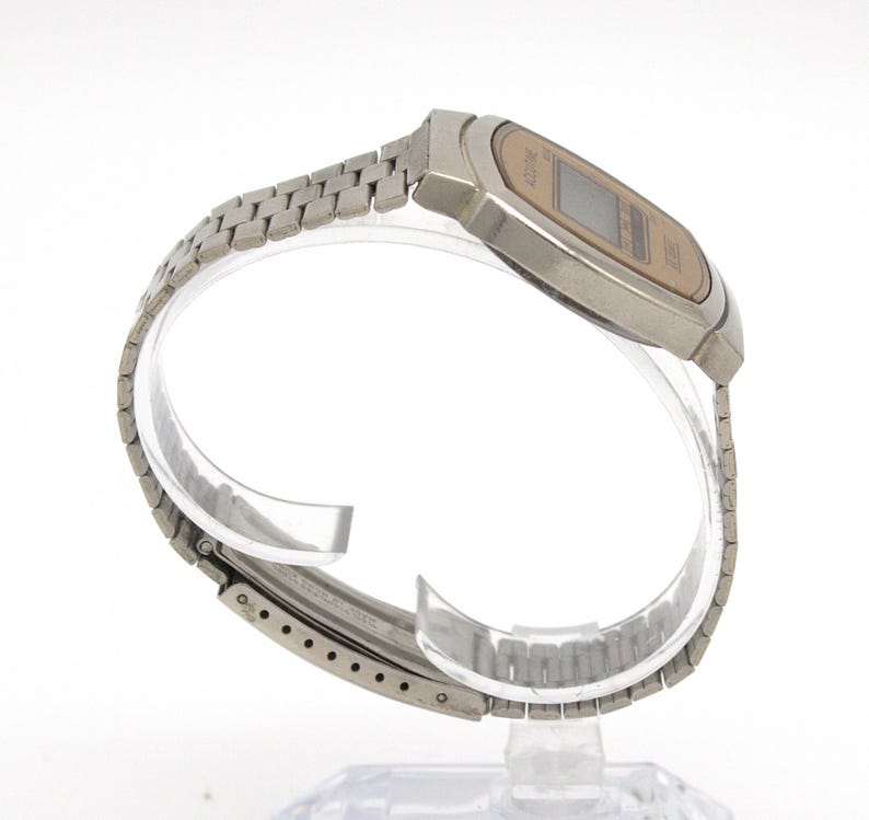 May include: A silver-toned digital watch with a rectangular face and a matching metal band. The watch face is a light tan color with a digital display. The band is made of small, connected metal links.