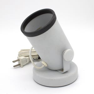 May include: A gray spotlight with a black rim and a white base. The lamp has a cylindrical shape and a white power cord with a two-prong plug. The lamp is angled upwards.