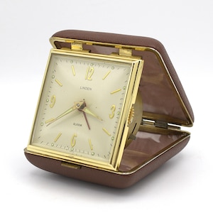 May include: A vintage Linden travel alarm clock in a brown leatherette case. The clock face is cream-colored with gold-tone hands and numerals. The case is open, revealing the clock's face and mechanism. The word "ALARM" is visible.