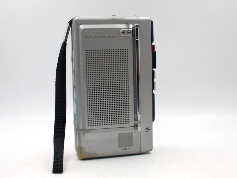May include: A silver and gray Sony radio cassette recorder with a black carrying strap. The radio has a telescoping antenna and a speaker grill. The text "SONY" and "RADIO CASSETTE-CORDER" are visible on the front.