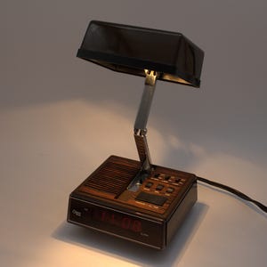 May include: A brown and black digital alarm clock with a built-in adjustable lamp. The base features a wood-grain finish, a speaker grille, and a red digital display showing 11:08. A chrome-colored metal arm connects the base to the rectangular lamp.