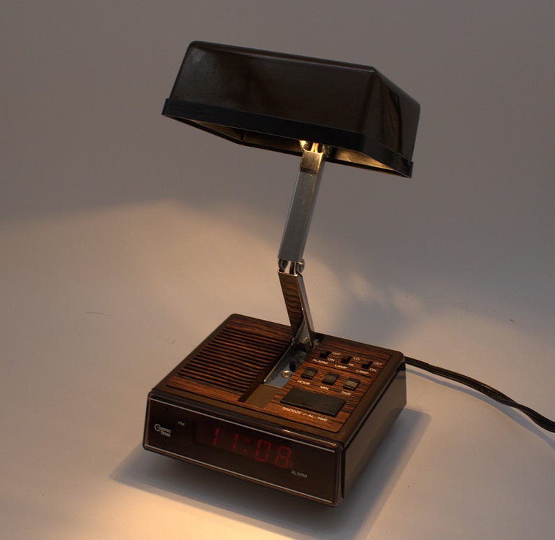 May include: A brown and black digital alarm clock with a built-in adjustable lamp. The base features a wood-grain finish, a speaker grille, and a red digital display showing 11:08. A chrome-colored metal arm connects the base to the rectangular lamp.