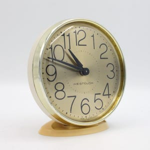 May include: A gold-colored Westclox alarm clock with black numbers and hands. The clock has a round face and a base.