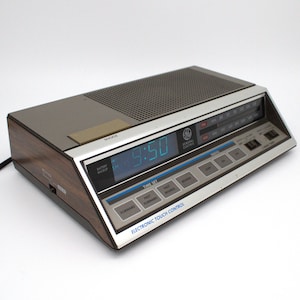 May include: A vintage General Electric digital alarm clock with a wood grain finish. The clock displays 9:50 in blue digits. Features include an AM/FM radio, snooze button, and electronic touch control.