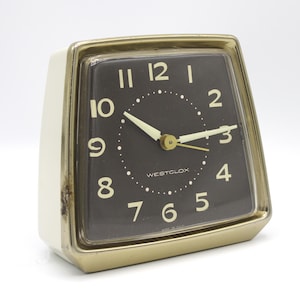 May include: Vintage Westclox alarm clock with a black face, gold-tone trim, and a cream-colored body. The clock features white numerals and hands, with the brand name "WESTCLOX" printed on the face. The clock is made in the USA.