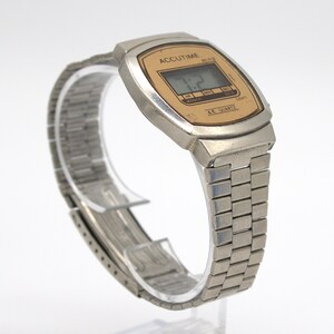 May include: A vintage Accutime digital watch with a silver-tone metal case and band. The watch face is rectangular with a gold-colored background and a digital display showing the time. The watch band is a flexible, linked design.
