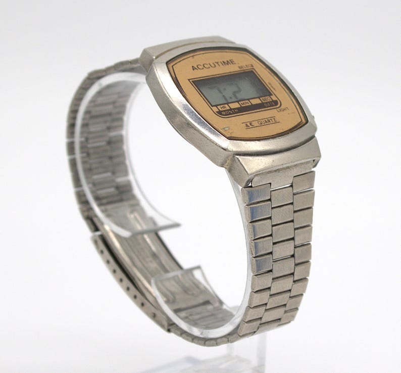 May include: A vintage Accutime digital watch with a silver-tone metal case and band. The watch face is rectangular with a gold-colored background and a digital display showing the time. The watch band is a flexible, linked design.