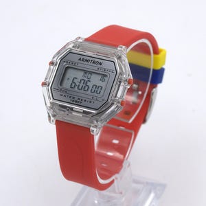 May include: A digital Armitron watch with a clear, octagonal case and a red silicone band. The watch face displays the time as 6:06:00. The band has a section with yellow, blue, and red stripes. The watch is water resistant to 100 feet.