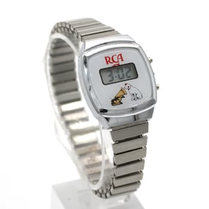 May include: A silver-tone digital wristwatch with a flexible metal expansion band. The white watch face features the red RCA logo, a digital time display showing 3:02, and a graphic illustration of the Nipper dog listening to a gramophone.