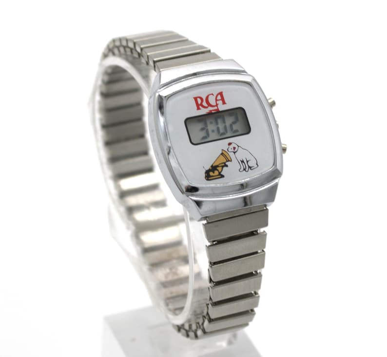 May include: A silver-tone digital wristwatch with a flexible metal expansion band. The white watch face features the red RCA logo, a digital time display showing 3:02, and a graphic illustration of the Nipper dog listening to a gramophone.