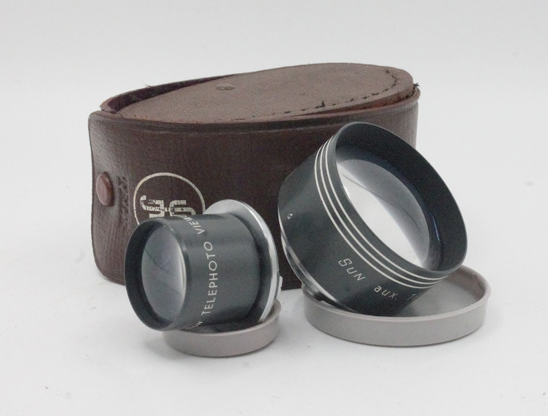 May include: Two vintage camera lens attachments in a brown leather case. The lenses are black with silver accents and have the text "Telephoto View" and "Aux Sun" printed on them.