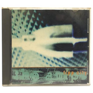 May include: A CD case with a teal, white, and blue abstract design. The cover features a blurred, elongated shape and a textured background. The text "Trip City" is visible at the bottom in teal and orange.