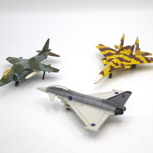 May include: Three toy airplanes are displayed against a white background. One is olive green and gray, another is yellow and brown camouflage, and the third is gray and white. The planes have detailed features.