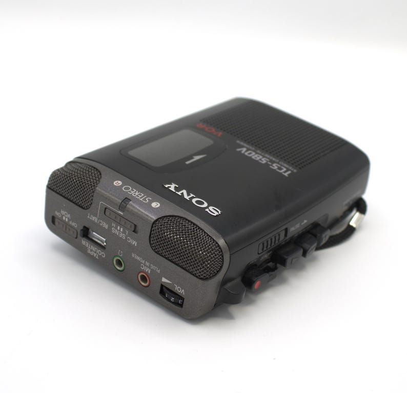 May include: A black Sony TCS-580V voice recorder with a dark gray front panel. The recorder has two built-in microphones, a volume control, and various input/output jacks. The word "SONY" is printed in white on the front.
