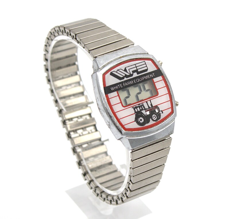 Peut inclure: Montre num&eacute;rique argent&eacute;e avec un cadran blanc et rouge. Le cadran affiche le texte "WHITE FARM EQUIPMENT" et un dessin de tracteur. La montre a un bracelet extensible argent&eacute;.