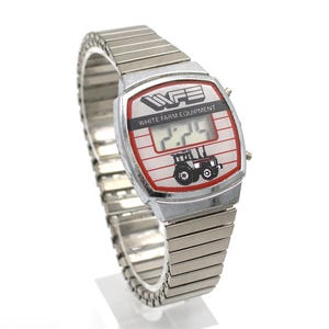 Peut inclure: Montre num&eacute;rique argent&eacute;e avec un cadran rectangulaire. Le cadran affiche le texte "WFE WHITE FARM EQUIPMENT" et un dessin de tracteur. L'heure affich&eacute;e est 2:24. Le bracelet est en m&eacute;tal argent&eacute; flexible. La montre mesure environ 3 cm de large.